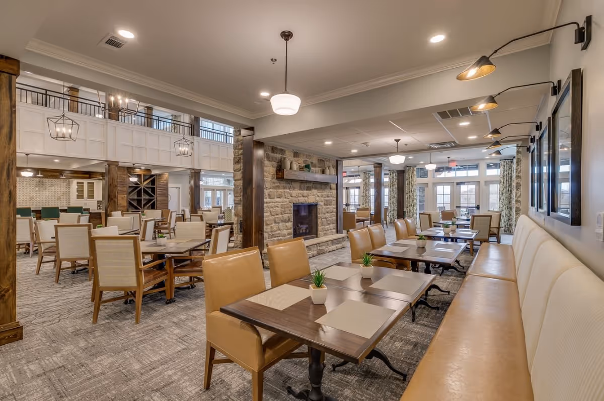 A spacious dining room in a senior living facility featuring multiple tables with beige placemats and small potted plants. The room has a stone fireplace in the center, leather chairs, and a long cushioned bench along one wall. Large windows with patterned curtains allow natural light to fill the space, and modern light fixtures hang from the ceiling.