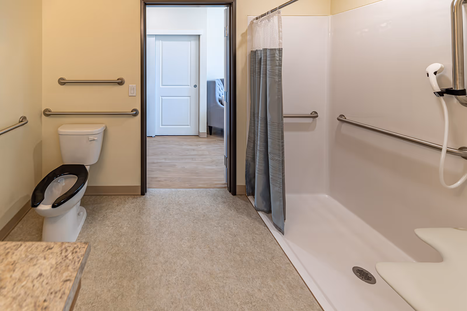 A clean and accessible bathroom featuring a toilet with a black seat, grab bars on the walls, and a walk-in shower with a shower curtain and handheld showerhead. The bathroom opens into a room with wooden flooring and a gray upholstered chair visible through the doorway.