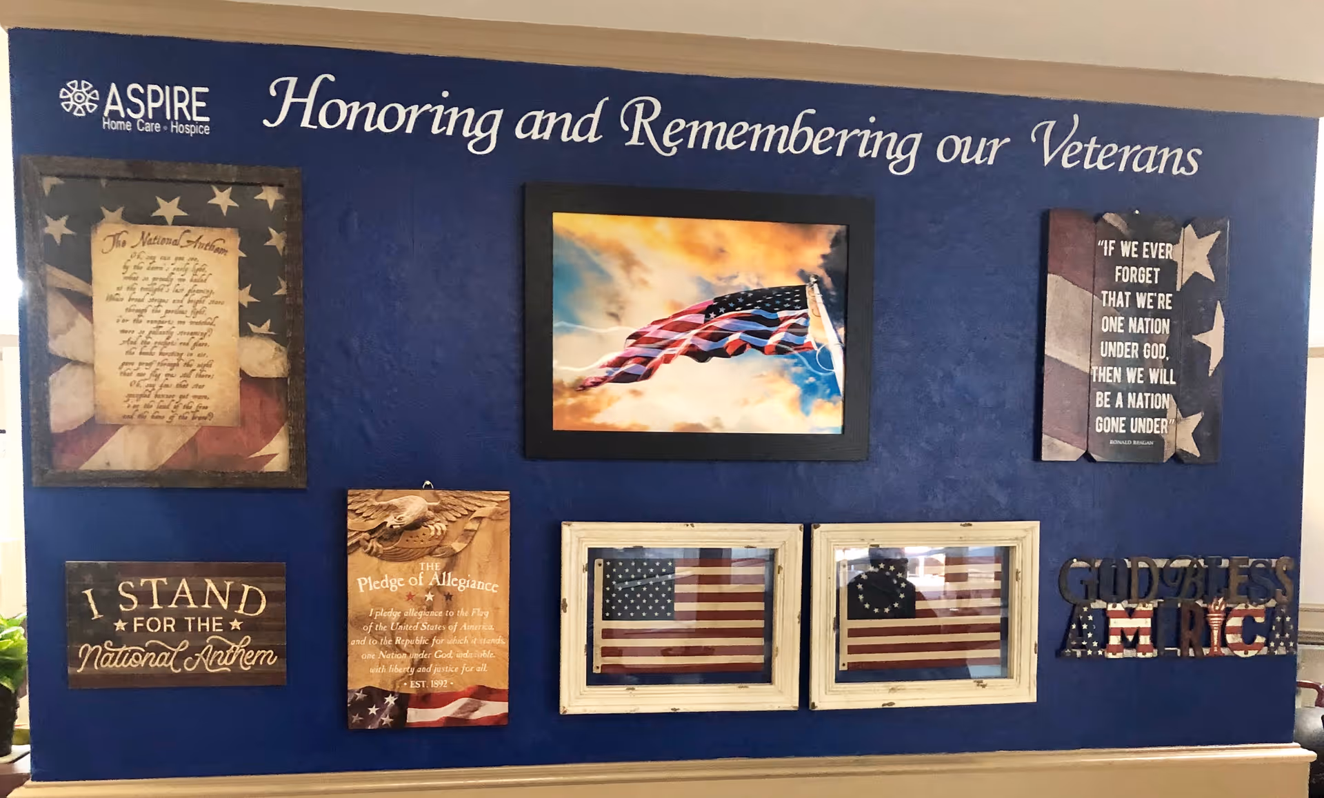 A blue wall display honoring and remembering veterans with various patriotic decorations including framed American flags, a photo of a waving American flag, plaques with the National Anthem and Pledge of Allegiance, a quote about unity under God, and a decorative sign saying 'God Bless America'.