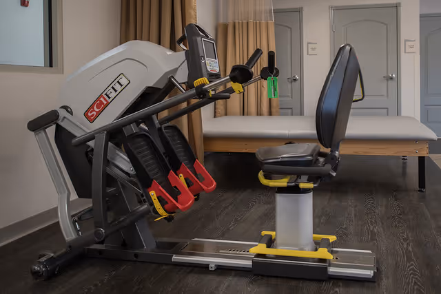 A rehabilitation exercise machine with foot pedals and handles, positioned in a room with wood flooring, beige curtains, and a padded examination table in the background.