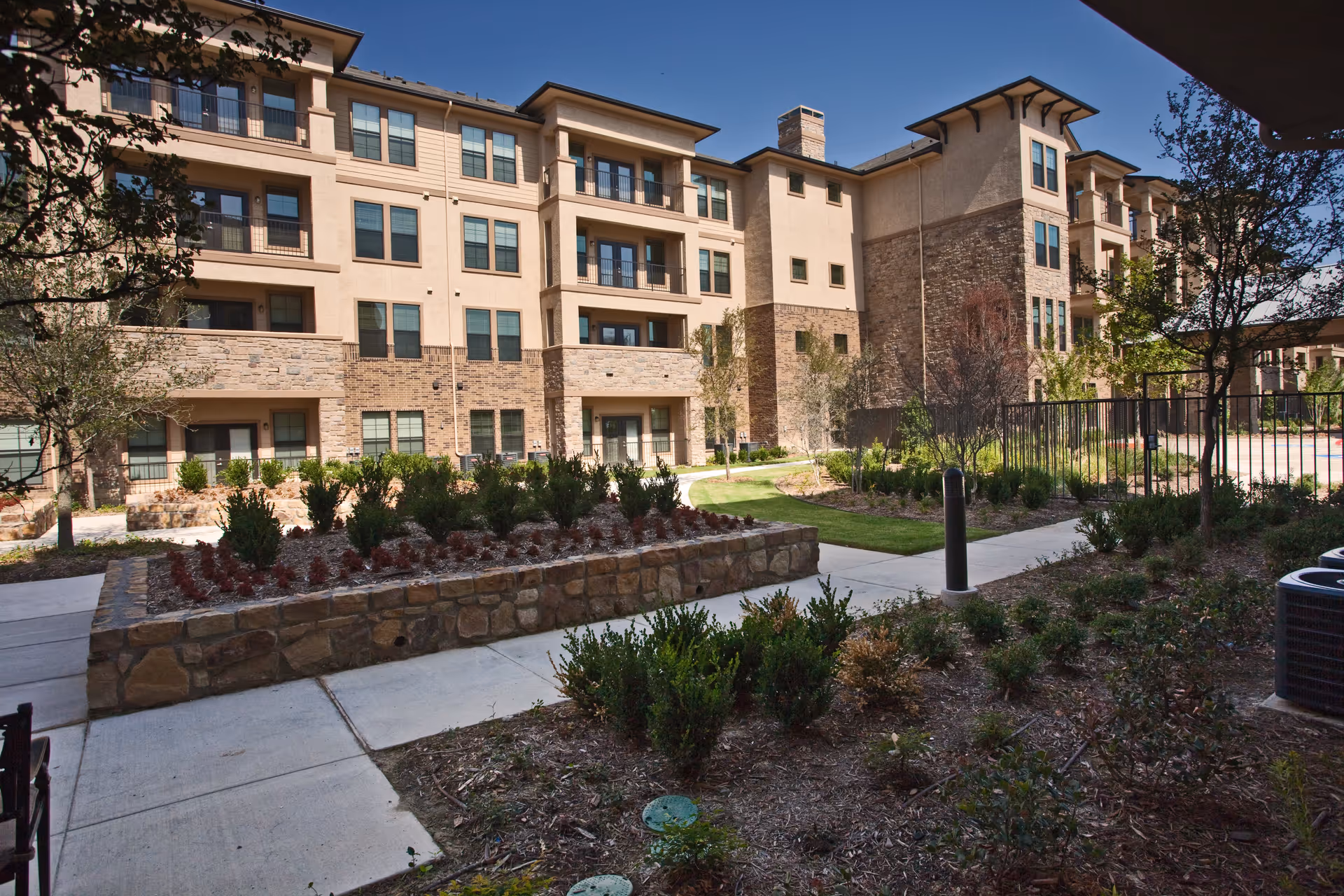 Exterior view of a multi-story senior living facility building with stone and beige siding, surrounded by landscaped gardens, shrubs, trees, and paved walkways under a clear blue sky.