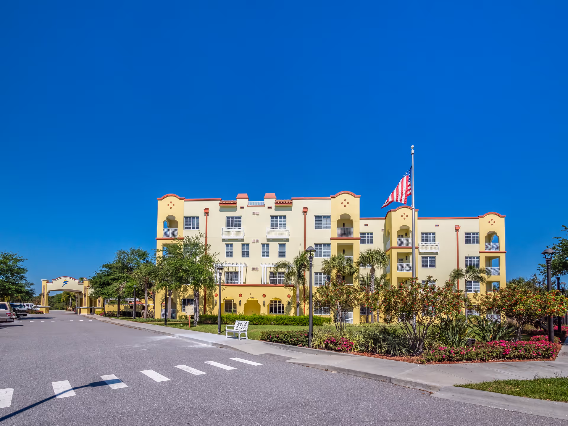 Exterior view of a multi-story yellow building with red trim, surrounded by palm trees and landscaped bushes. An American flag is flying on a flagpole in front of the building. There is a paved road with a pedestrian crosswalk and a white bench near the sidewalk. The sky is clear and blue.