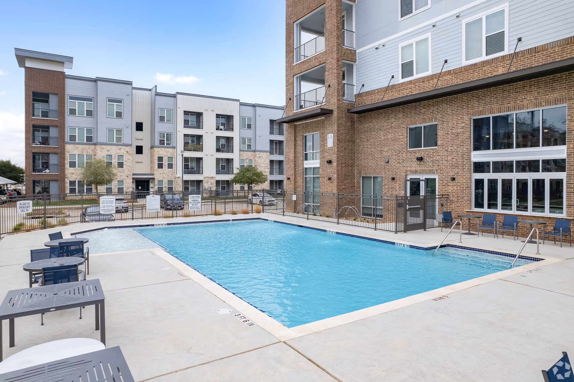 Outdoor swimming pool area surrounded by a concrete deck with lounge chairs and tables, adjacent to a multi-story residential building with balconies and large windows under a partly cloudy sky.
