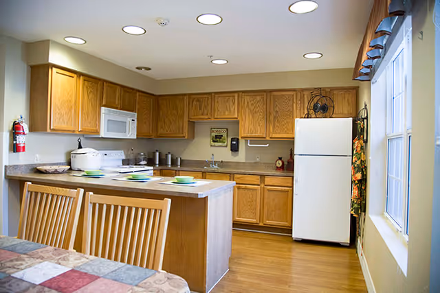 A kitchen with wooden cabinets, a white refrigerator, a microwave, a stove, and a countertop with three place settings. There is a window with a valance on the right side and a fire extinguisher mounted on the wall to the left.