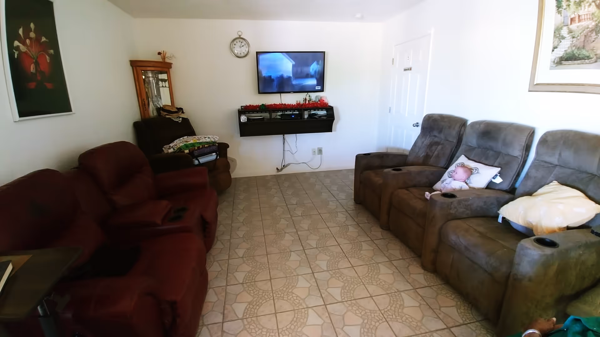 A cozy living room with two sets of reclining chairs facing a wall-mounted flat screen TV. The floor is tiled with a patterned design. On the left side, there is a wooden cabinet with glass doors and a painting of flowers on the wall. On the right side, there is a framed picture hanging on the wall. A doll is placed on one of the reclining chairs. A clock is mounted on the wall above the TV.