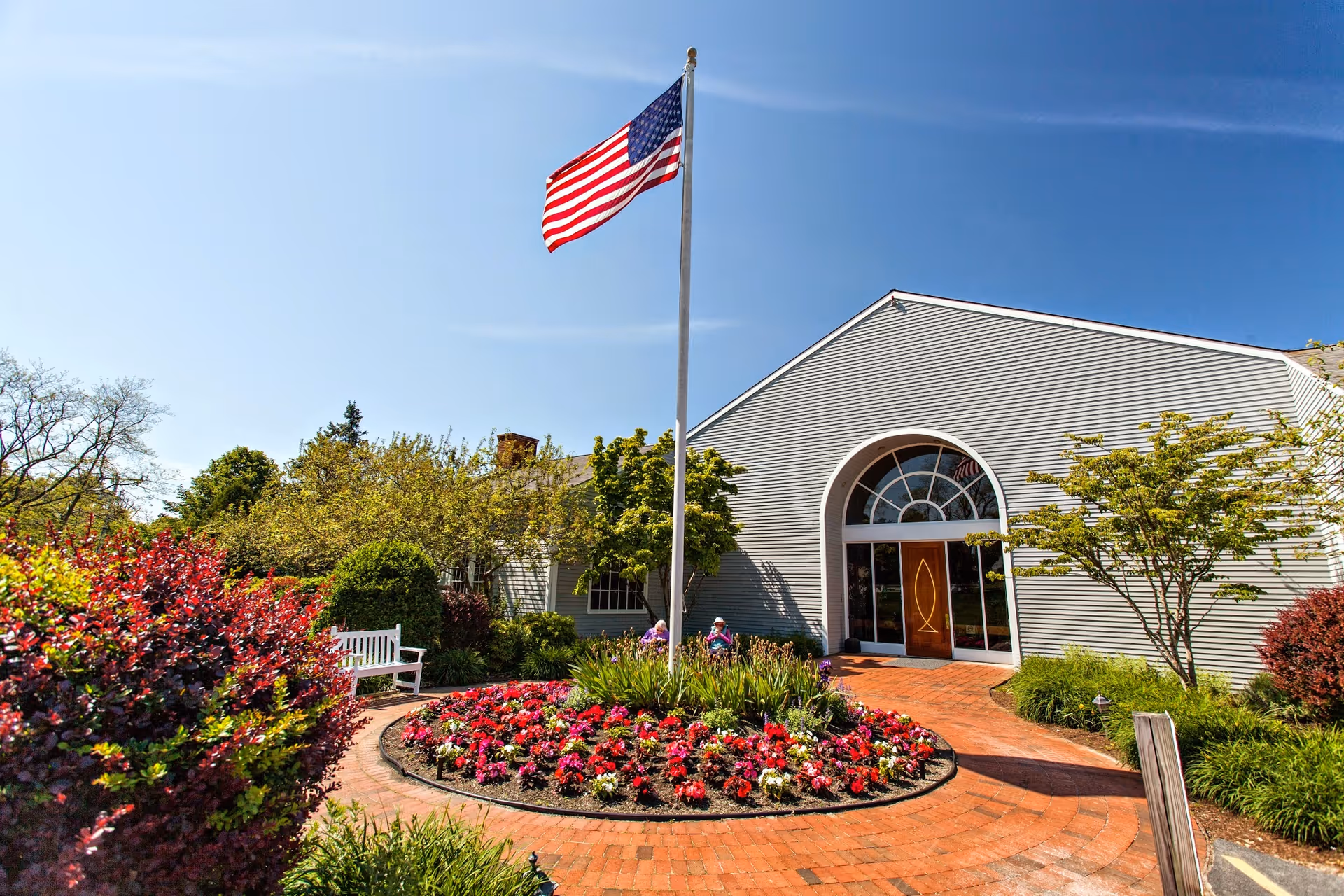 Front entrance of a healthcare building with an American flag, circular flower bed and brick walkway leading to an arched double door.