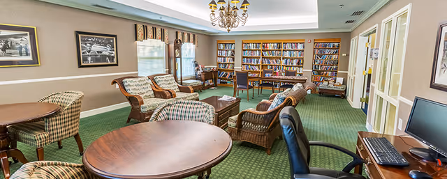 A cozy common room with green carpet and beige walls, featuring several wooden tables and cushioned chairs with plaid upholstery. There are framed black and white photographs on the walls, a grandfather clock near the windows, and a large bookshelf filled with books at the far end of the room. A computer workstation with a monitor and keyboard is visible on the right side.
