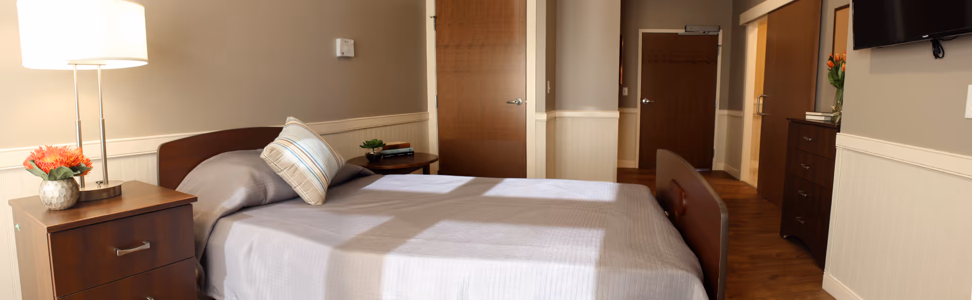 Well-lit senior bedroom with a made bed, nightstand and lamp, dresser with TV, and doors leading to a hallway.