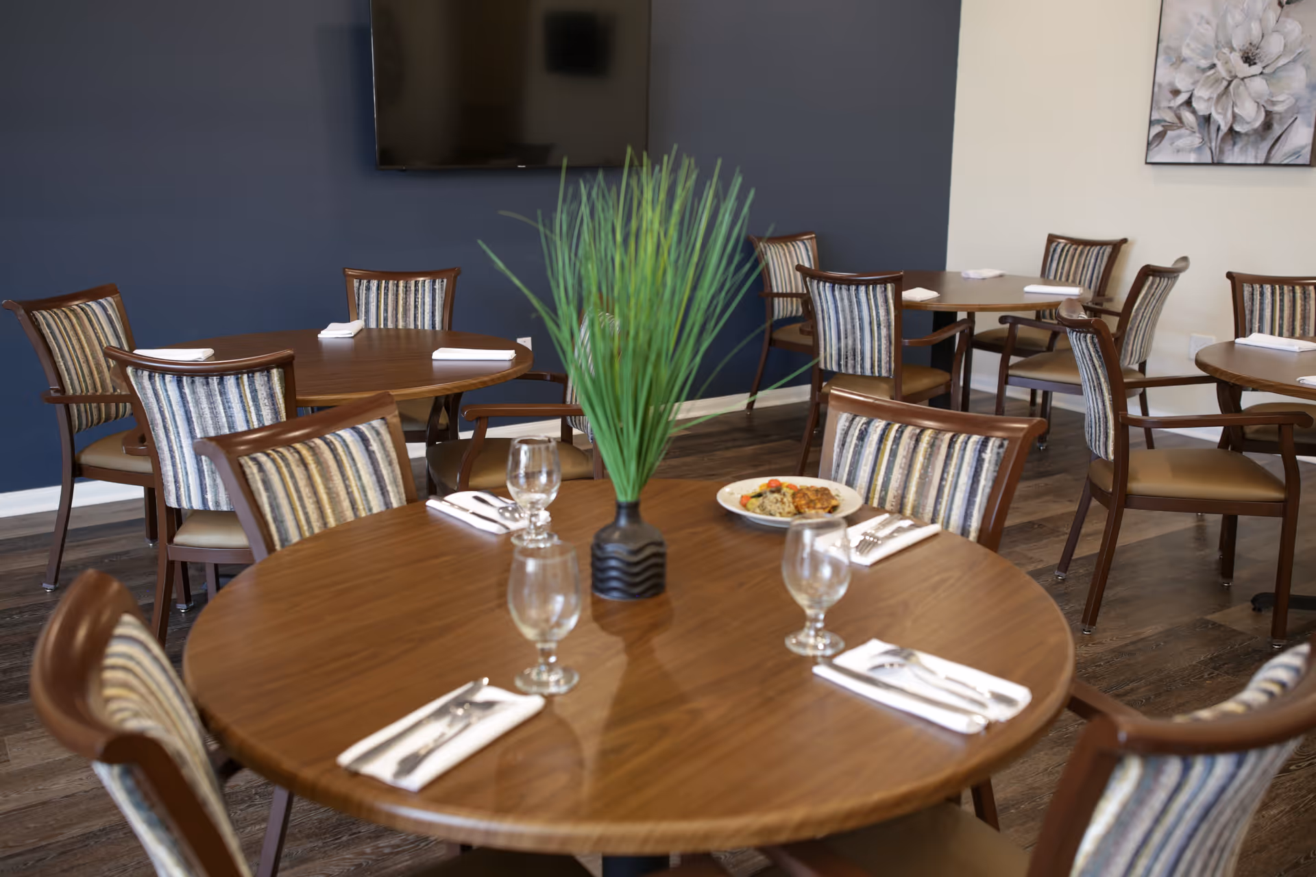 Dining room with round wooden tables and striped cushioned chairs. Tables are set with napkins, silverware, and empty glasses. One table has a small black vase with green grass-like plant and a plate of food. The room has dark wood flooring, a dark blue accent wall with a mounted TV, and a floral painting on a light-colored wall.