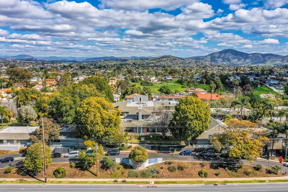 Aerial view of Monte Vista Village senior living facility surrounded by trees and greenery, with a suburban neighborhood and mountains in the background under a partly cloudy sky.