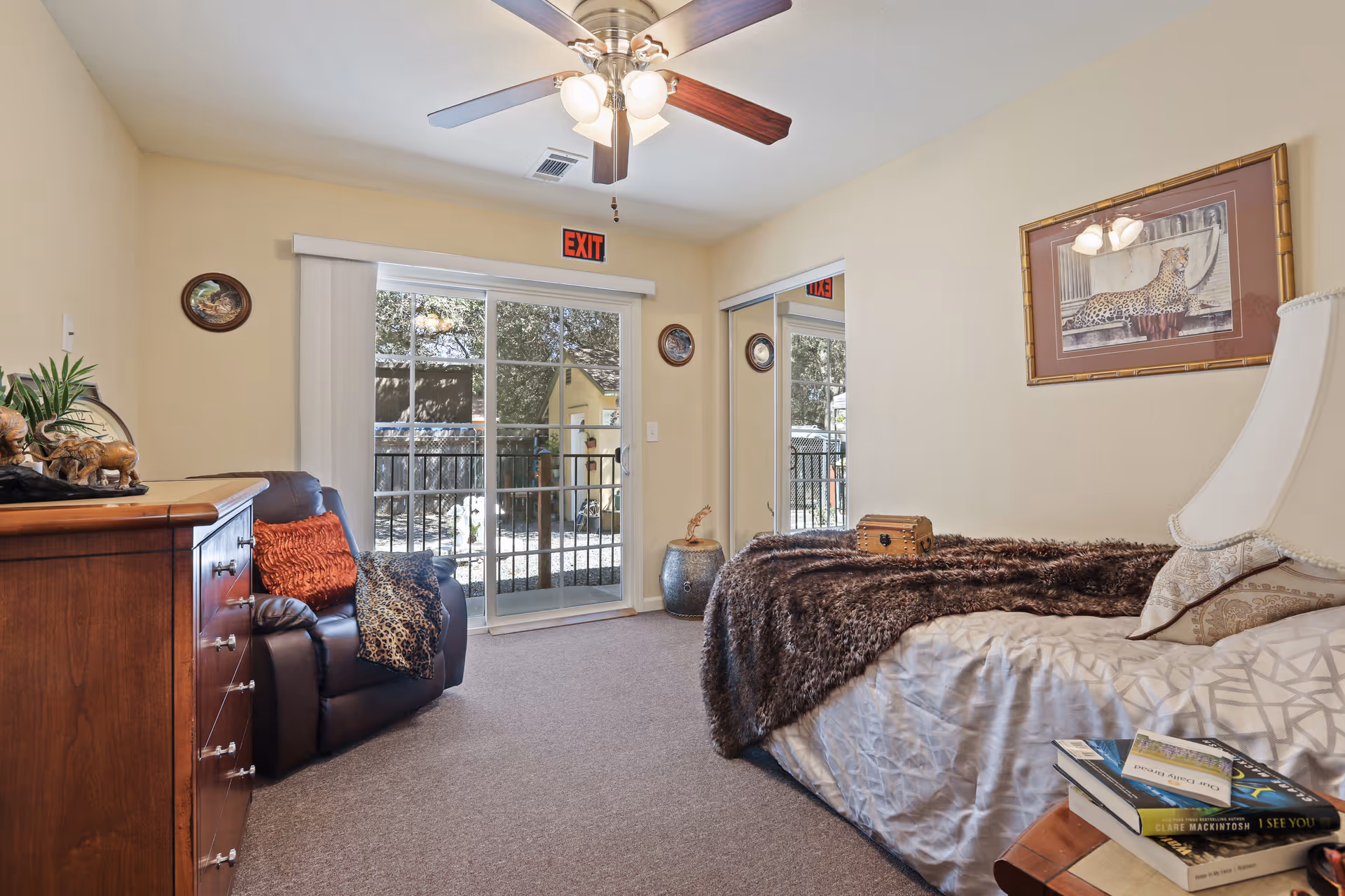 A cozy bedroom in a senior living facility featuring a bed with a brown fur blanket, a bedside table with books, a lamp, a leather armchair with decorative pillows, a wooden dresser, and a ceiling fan. There is a sliding glass door leading outside and a mirrored closet door on the right wall. The walls are decorated with framed artwork and small round plates.