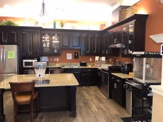 A modern kitchen with dark wood cabinets, granite countertops, and stainless steel appliances including a refrigerator and stove. There is a kitchen island with a wooden chair, a popcorn machine on the right side, and various kitchen utensils and appliances on the counters. The floor is wood, and the walls are painted a warm orange color.