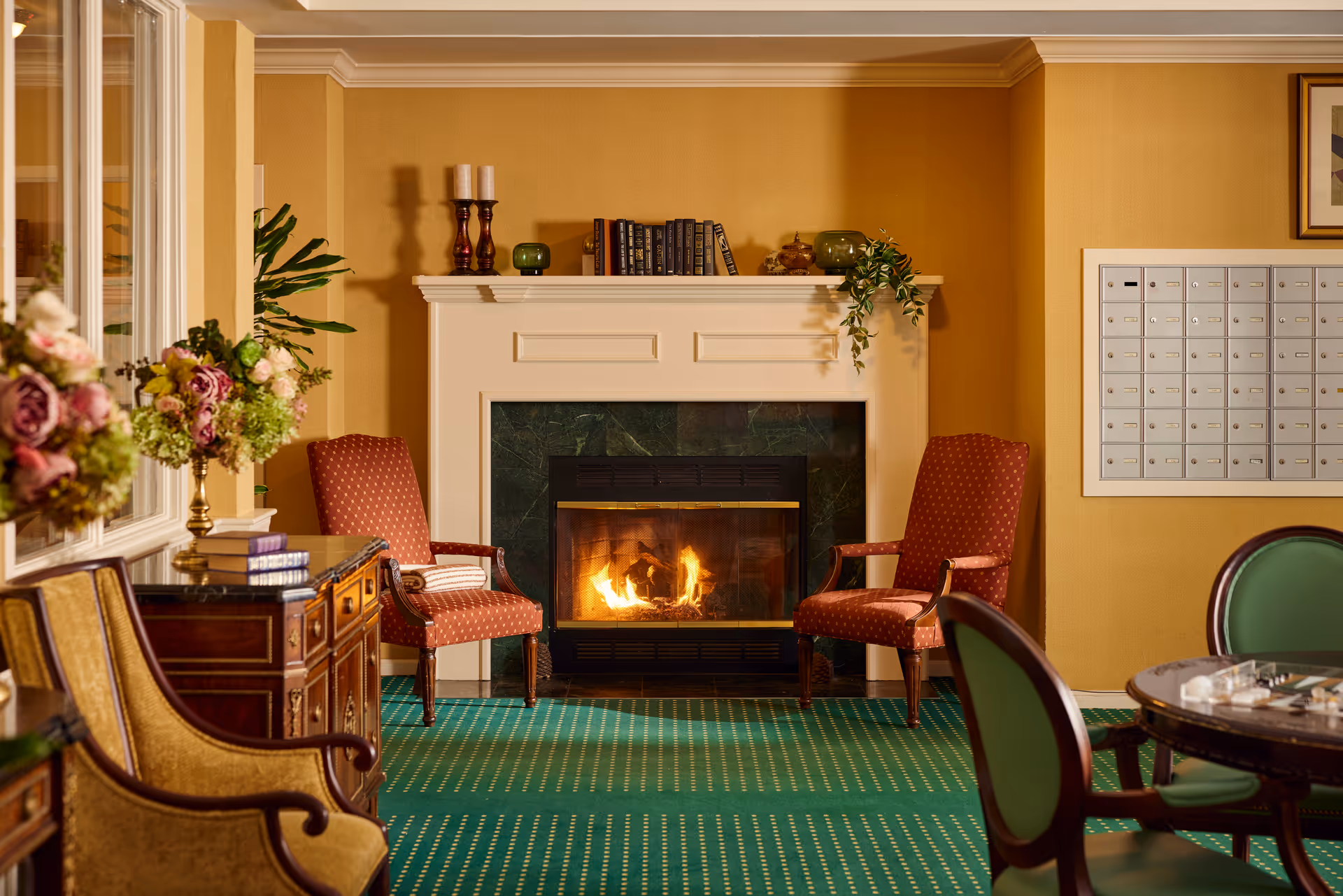 Cozy seating area with two upholstered armchairs flanking a lit fireplace, a decorated mantle, green carpet, and a wall of mailboxes.