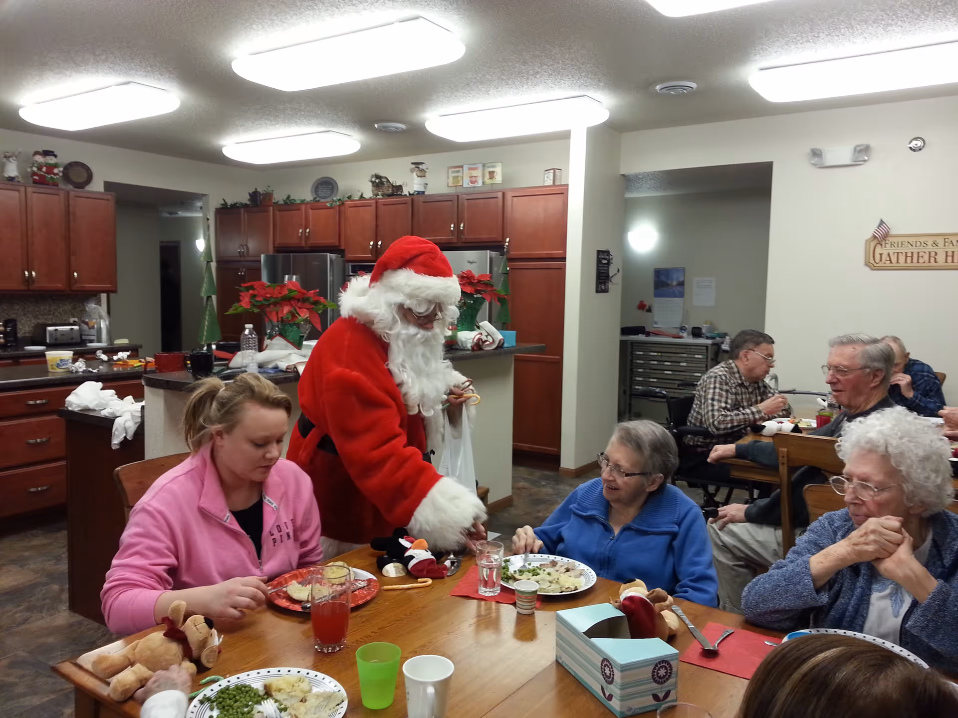 A group of elderly people and a younger woman sitting around a dining table in a kitchen area, having a meal. A person dressed as Santa Claus is standing and interacting with one of the elderly women. The kitchen has wooden cabinets, a refrigerator, and festive decorations including poinsettias and holiday figurines.