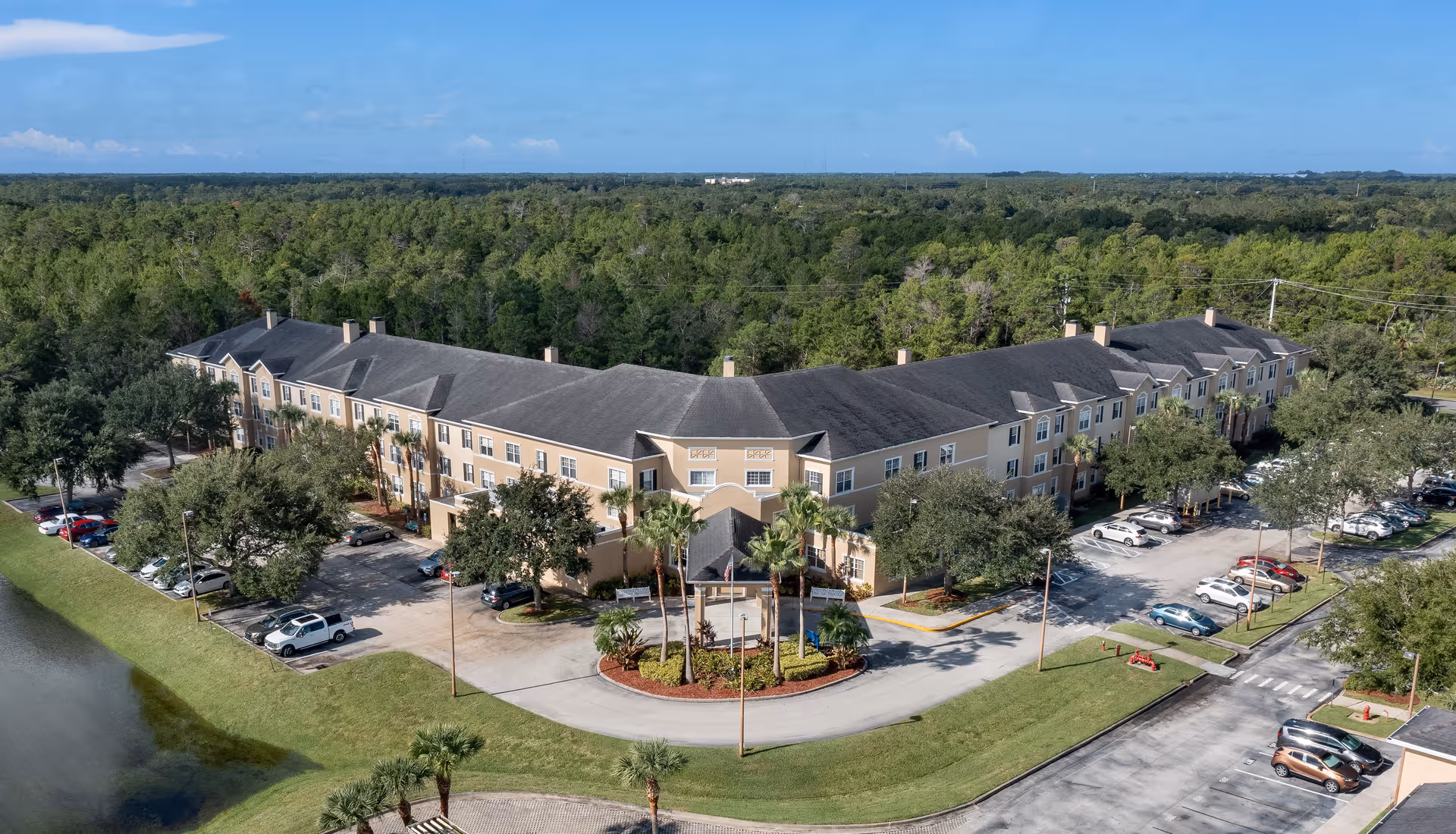 Aerial view of a large senior living facility building with a U-shaped layout surrounded by trees and greenery. The building has a beige exterior with multiple windows and a dark roof. There is a circular driveway with palm trees and landscaped greenery in front of the main entrance. Several cars are parked in parking lots on either side of the building. The background shows a dense forested area under a clear blue sky.