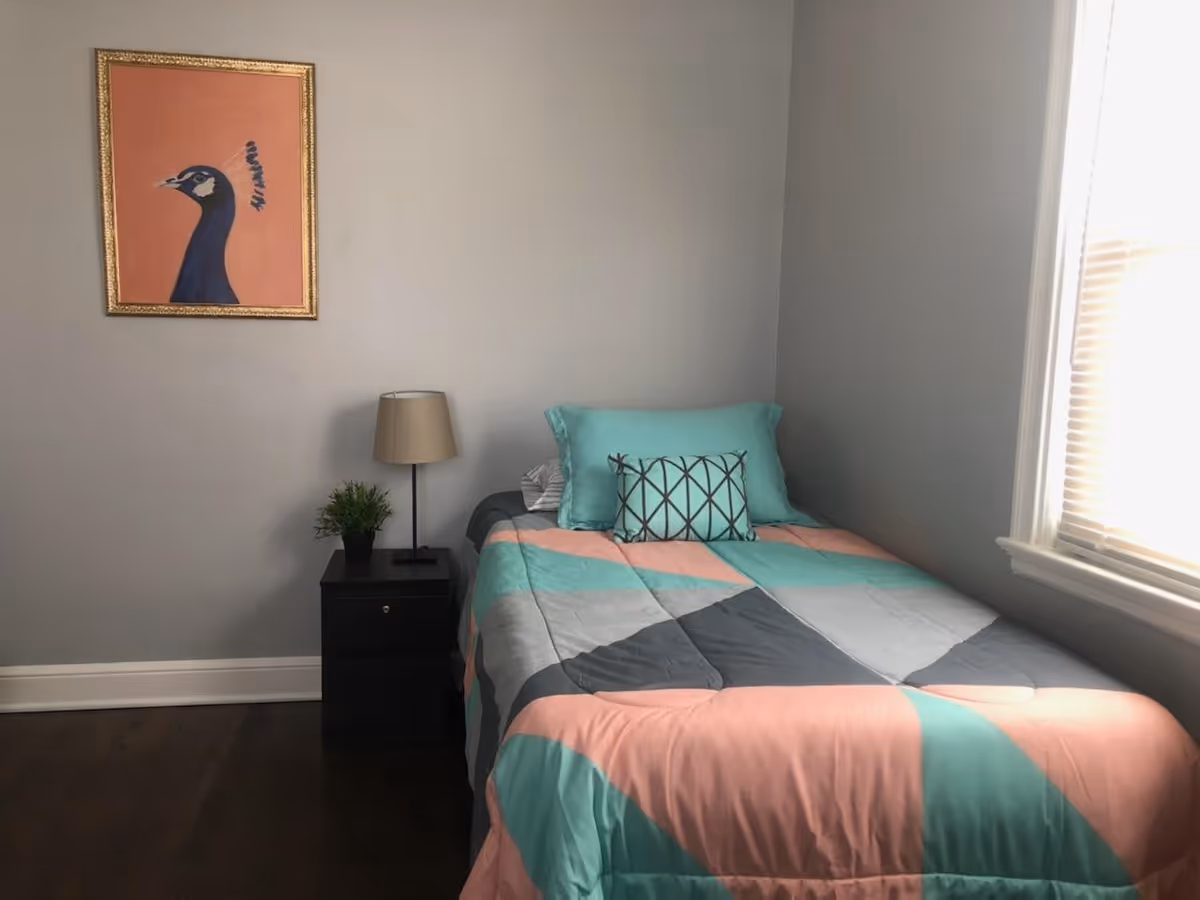 A small bedroom with a single bed covered in a colorful quilt with teal, pink, gray, and white geometric patterns. There are two pillows on the bed, one teal and one with a crisscross pattern. Next to the bed is a black nightstand with a small green plant and a beige lamp. A framed painting of a peacock on a peach background hangs on the gray wall above the nightstand. A window with closed blinds is on the right side of the room.
