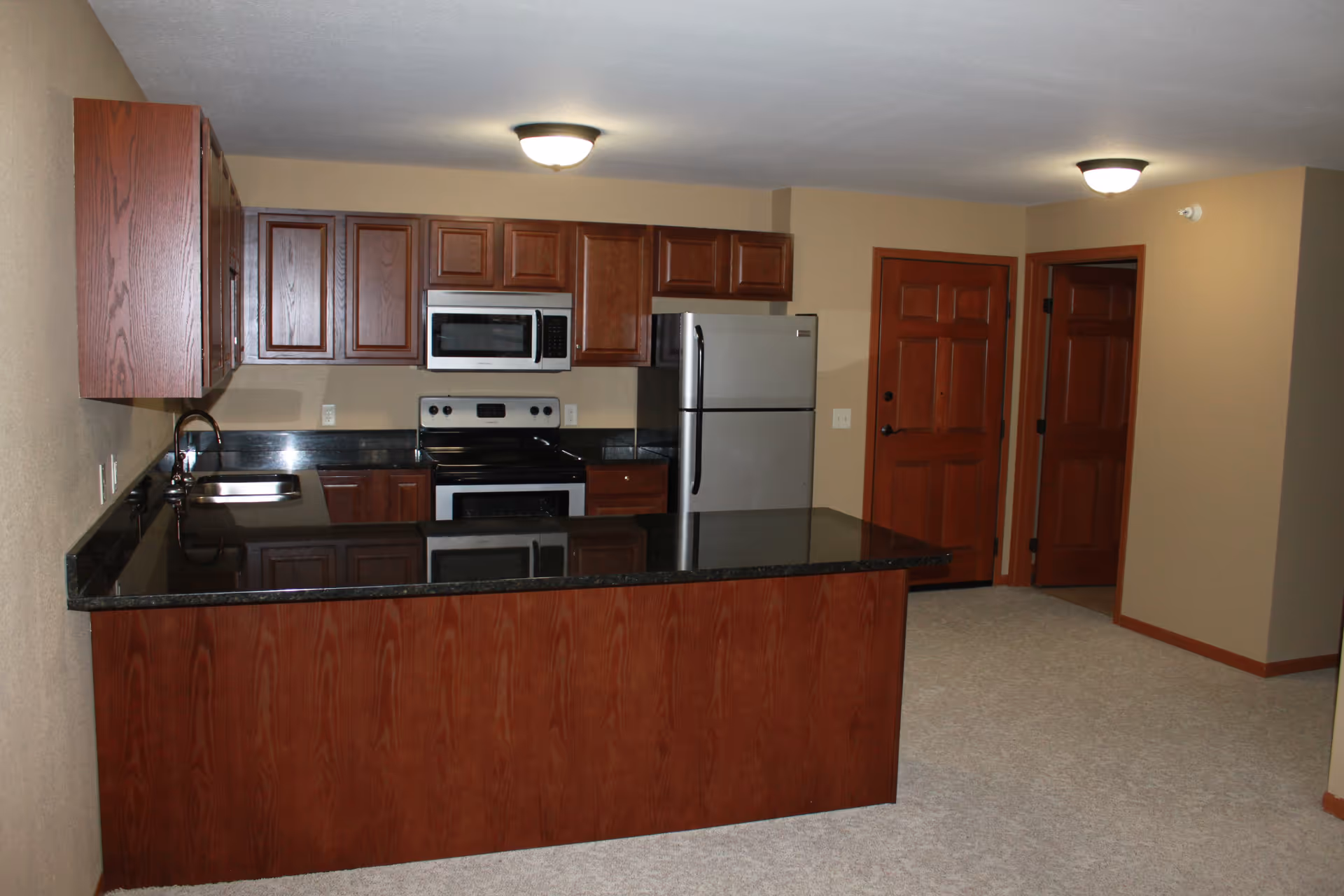 Open kitchen with a dark granite island, wooden cabinets, stainless steel appliances, and nearby entry doors.