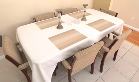 A dining table covered with a white tablecloth and four beige striped placemats. The table is surrounded by eight beige striped chairs with wooden legs. Two decorative candle holders and a small glass dish are placed in the center of the table. The room has tiled flooring and beige walls.