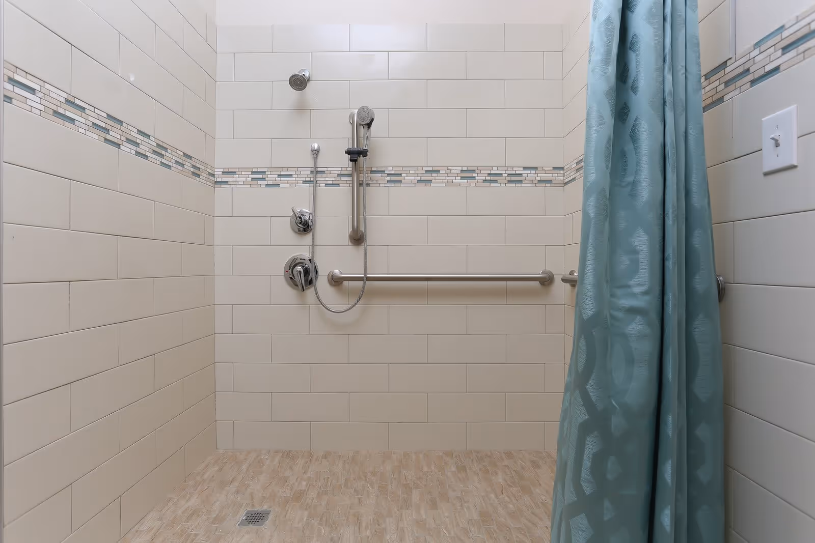 Tiled walk-in shower with a handheld showerhead, horizontal grab bar and teal shower curtain.