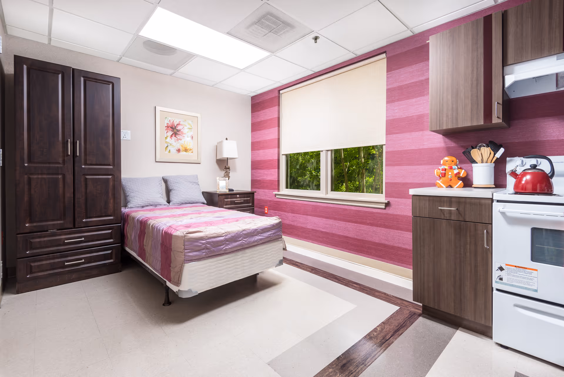 A small, cozy room with a single bed covered in a pink and purple striped bedspread, two gray pillows, a dark wooden wardrobe and nightstand with a lamp, a window with a beige roller shade, and a kitchenette area with a stove, cabinets, and a red kettle. The walls have a pink striped accent wall and a floral picture hanging above the bed.