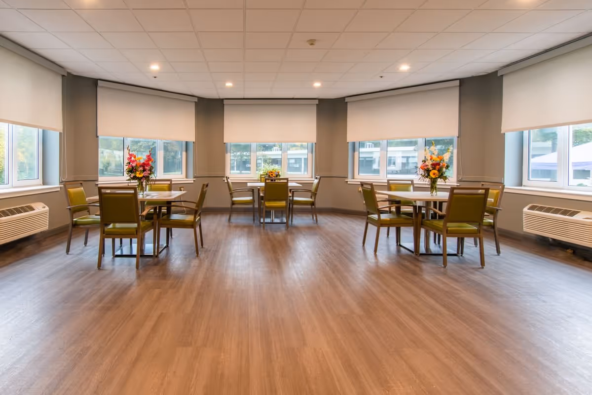 A bright dining room with three round tables, each surrounded by four green cushioned chairs. Each table has a vase with colorful flowers. Large windows with white roller blinds let in natural light, and there are air conditioning units below the windows. The floor is wooden and the ceiling has recessed lighting.