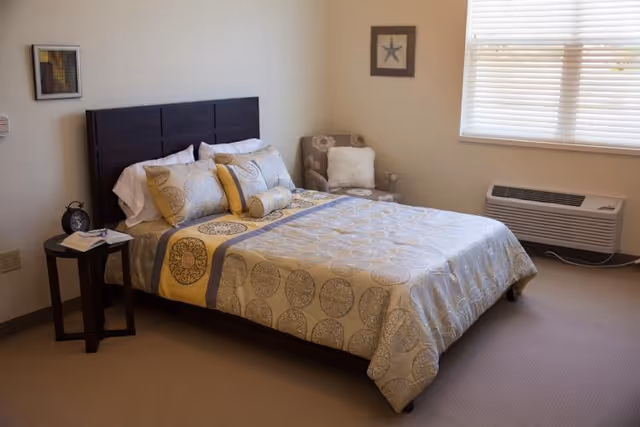 A neatly made bed with patterned bedding and multiple pillows in a bedroom. Next to the bed is a small round side table with a clock and a book. There is a cushioned armchair with a pillow in the corner near a window with blinds. An air conditioning unit is installed below the window. The walls are decorated with two framed pictures.