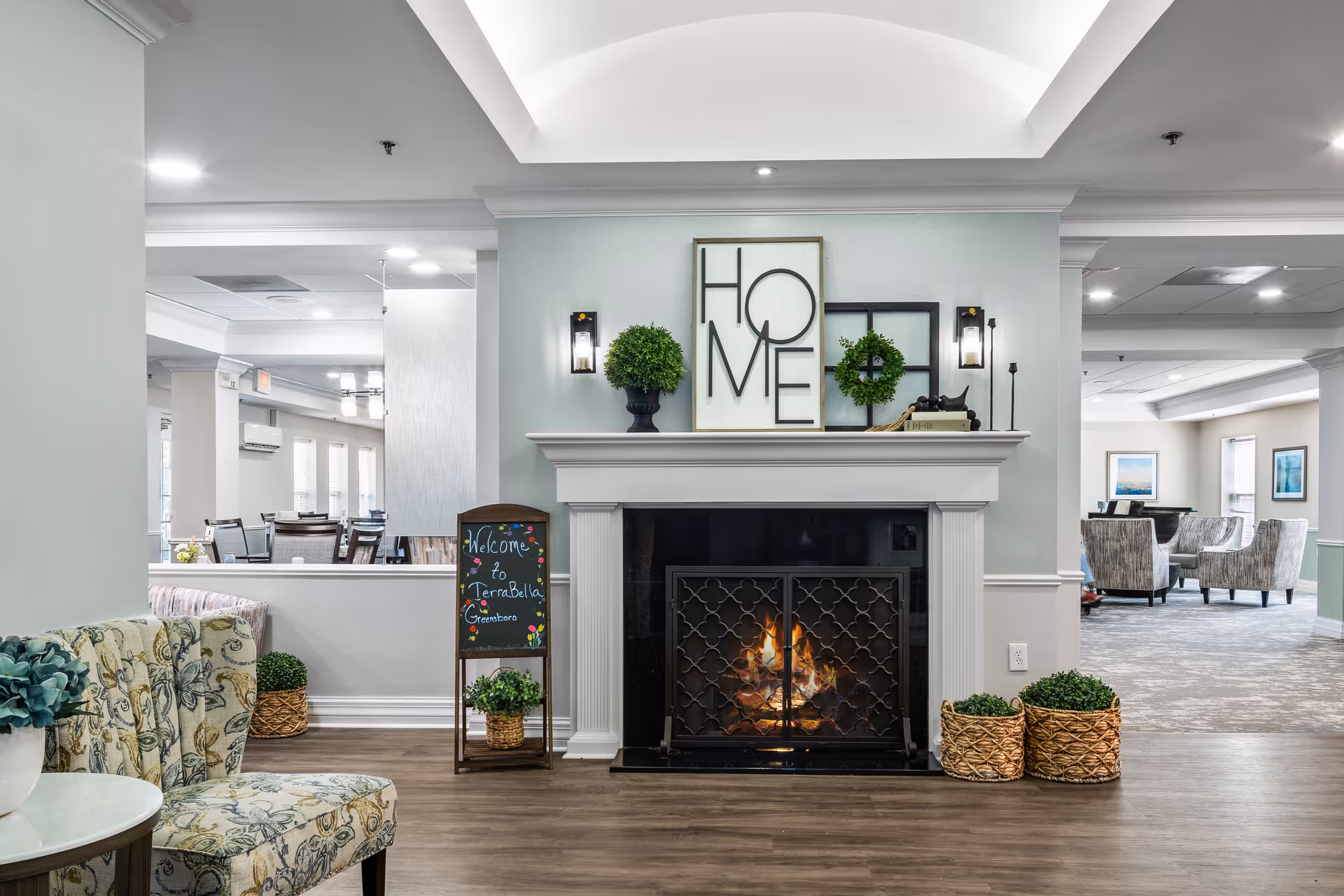 A cozy interior common area with a lit fireplace in the center, decorated with a framed 'HOME' sign, greenery, and a small chalkboard sign that says 'Welcome to TerraBella Greensboro'. There are patterned chairs and tables around, with soft lighting and a welcoming atmosphere.