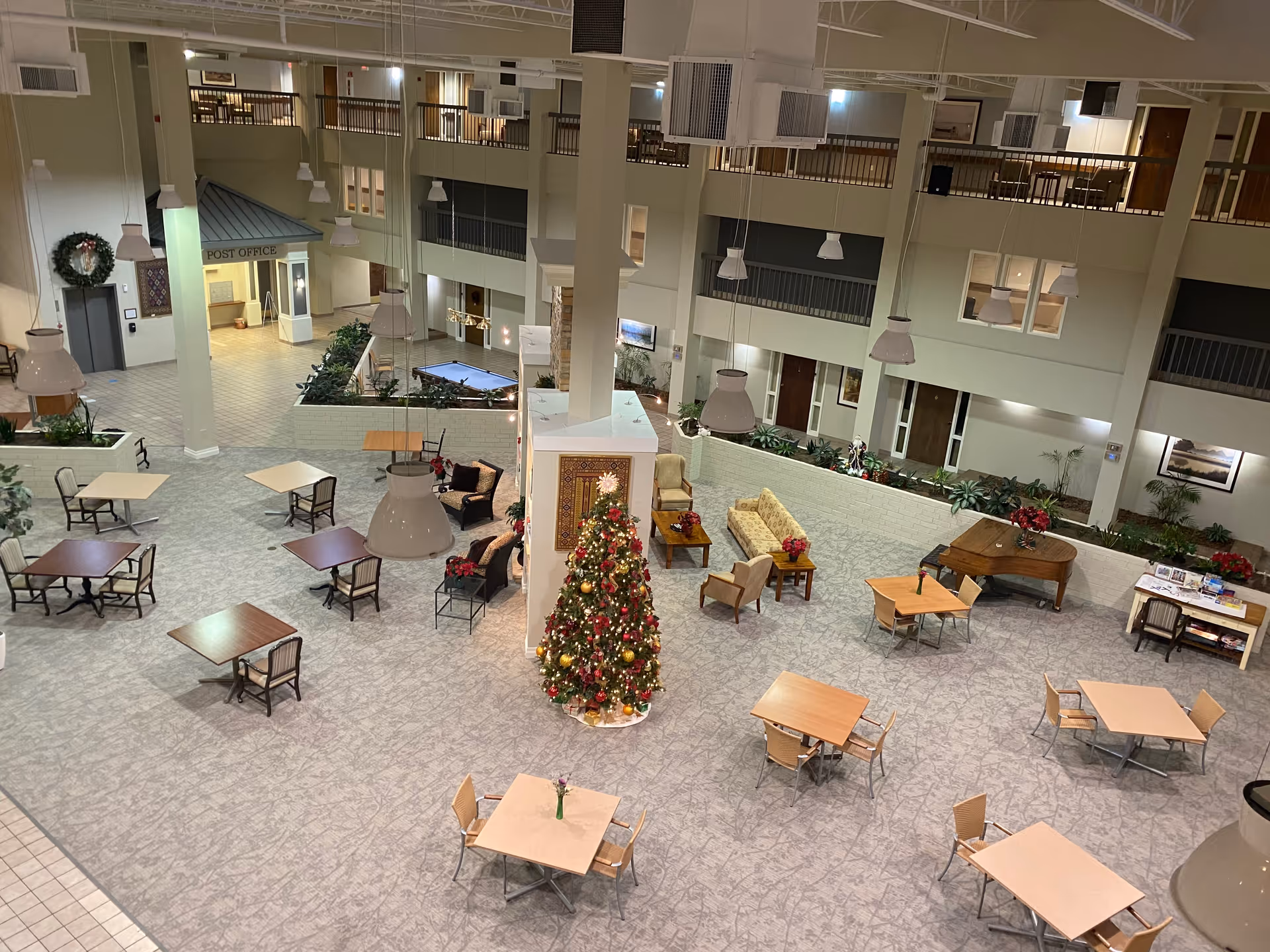 Spacious senior living facility common area decorated for Christmas with a large Christmas tree in the center. The area features multiple tables and chairs, a grand piano, comfortable seating arrangements, plants, and a post office section in the background. The space has high ceilings with hanging lights and a second-floor balcony overlooking the area.