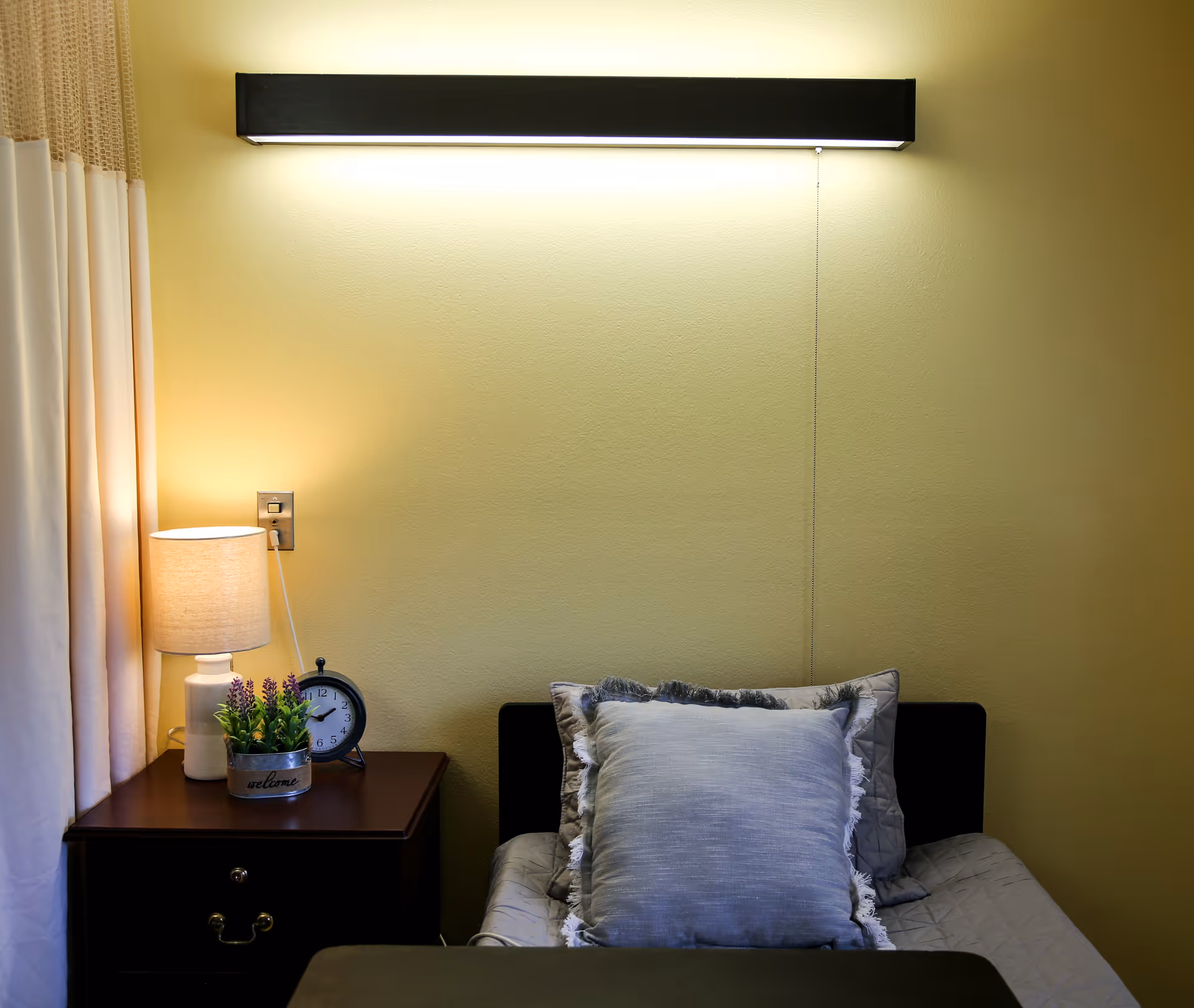 A cozy bedroom corner with a single bed featuring a gray pillow and quilt. Next to the bed is a dark wooden nightstand with a small lamp, a round black alarm clock, and a small potted plant with purple flowers. The wall behind the bed is painted light yellow, and a long rectangular light fixture is mounted above the bed. Cream-colored curtains are partially visible on the left side.