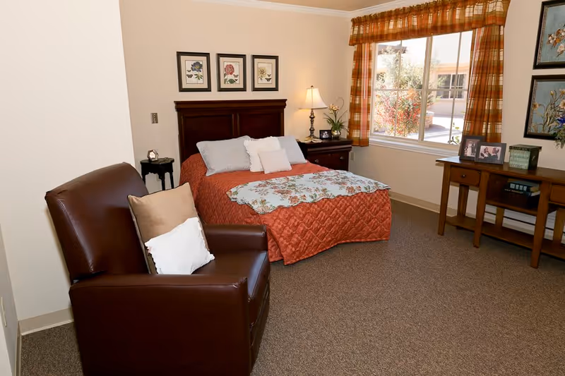 A cozy bedroom in a senior living facility featuring a bed with a red quilt and floral blanket, a brown leather armchair with pillows, a wooden nightstand with a lamp and flowers, a window with plaid curtains, and a wooden table with framed photos and decorative items.