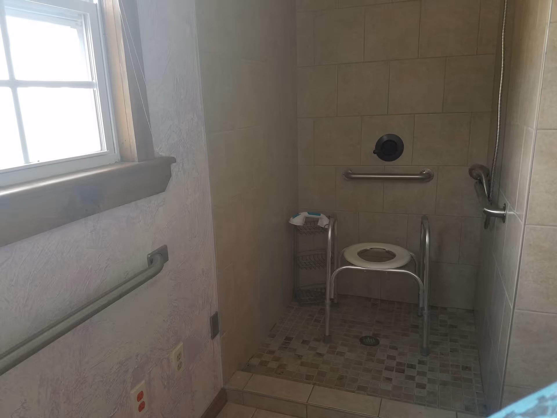 A tiled shower area with a shower chair, grab bars on the walls, and a small corner shelf. There is a window with wooden trim on the left side and a tiled floor with a drain in the center.