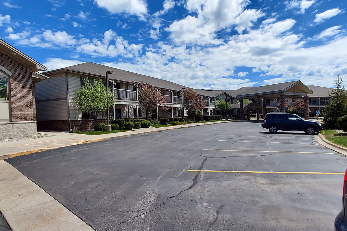 Exterior view of a senior living facility with a large parking lot in front, two-story buildings with balconies, trees, and a partly cloudy blue sky.