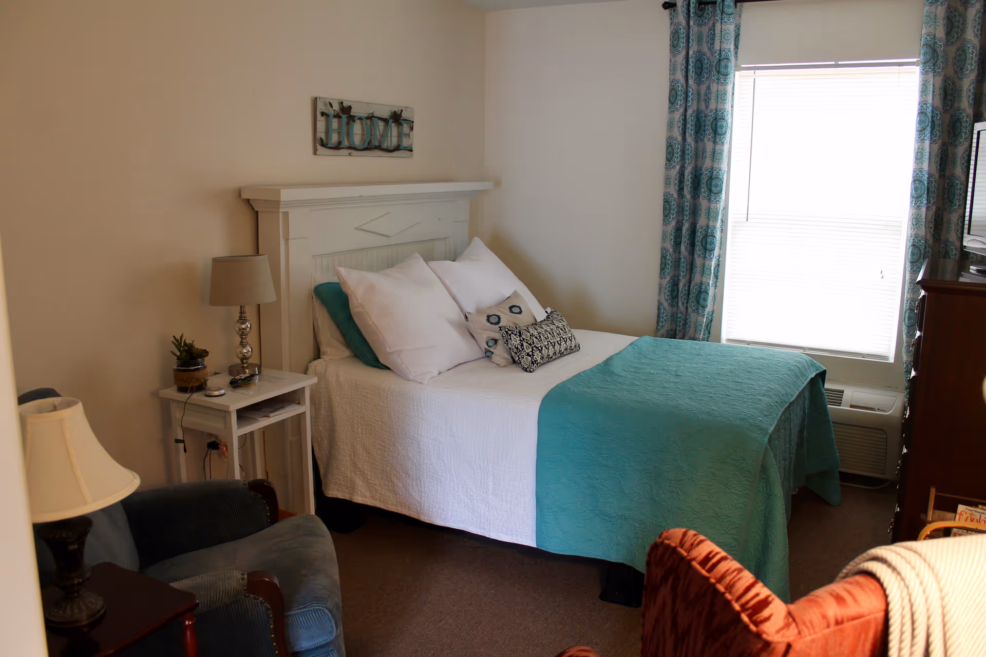 A cozy bedroom with a white bed featuring teal and patterned pillows and a teal blanket. There is a white nightstand with a lamp and small plant next to the bed. Two armchairs, one blue and one red, are positioned in the room. A window with teal patterned curtains lets in natural light. A decorative sign that says 'HOME' hangs above the bed's headboard.
