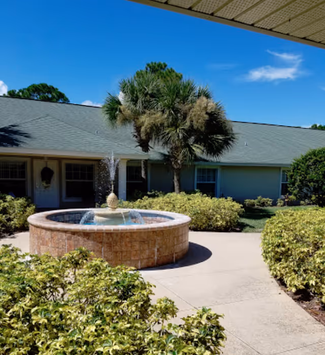 Outdoor courtyard area with a circular stone fountain in the center, surrounded by green bushes and a palm tree. A single-story building with a green roof and windows is visible in the background under a clear blue sky.