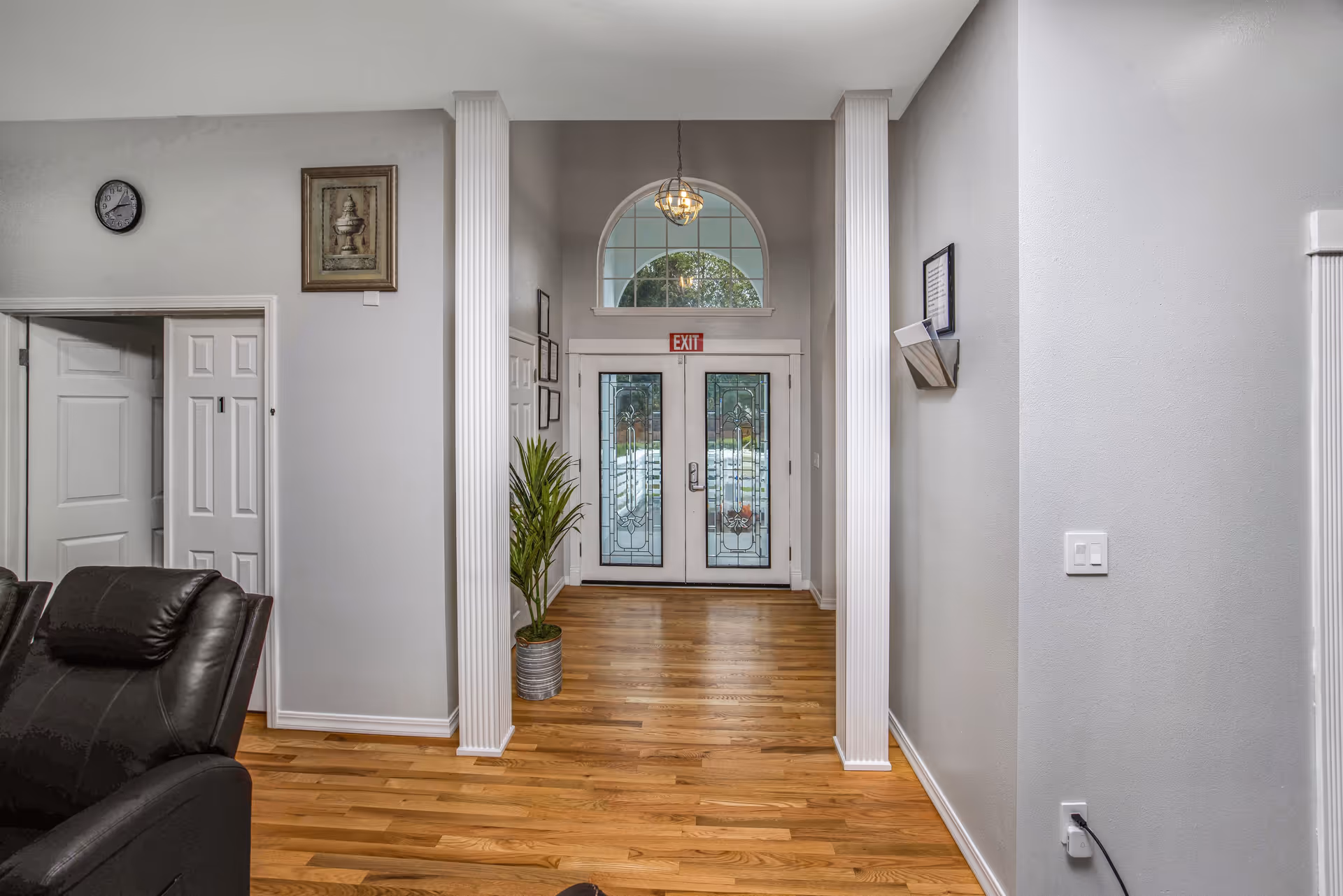 Interior view of a hallway with wooden flooring leading to a double glass door with decorative patterns. The hallway features two white columns, a hanging light fixture, a potted plant on the left side, a clock and framed artwork on the left wall, and a wall-mounted holder with papers on the right wall. Part of a black leather chair is visible in the foreground on the left.