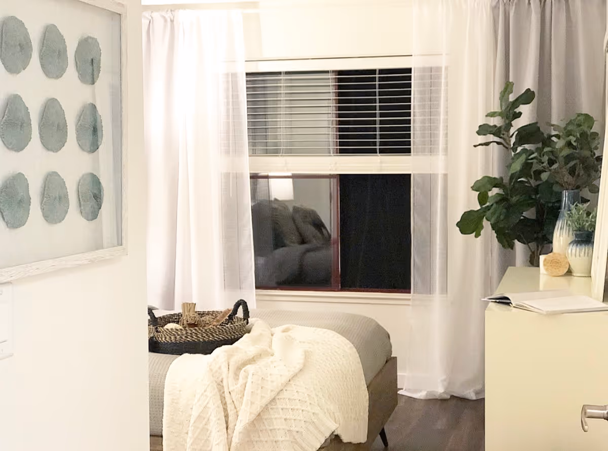 Cozy bedroom with a bed draped in a cream throw, sheer curtains over a window, a dresser topped with a potted plant and decor, and framed wall art.