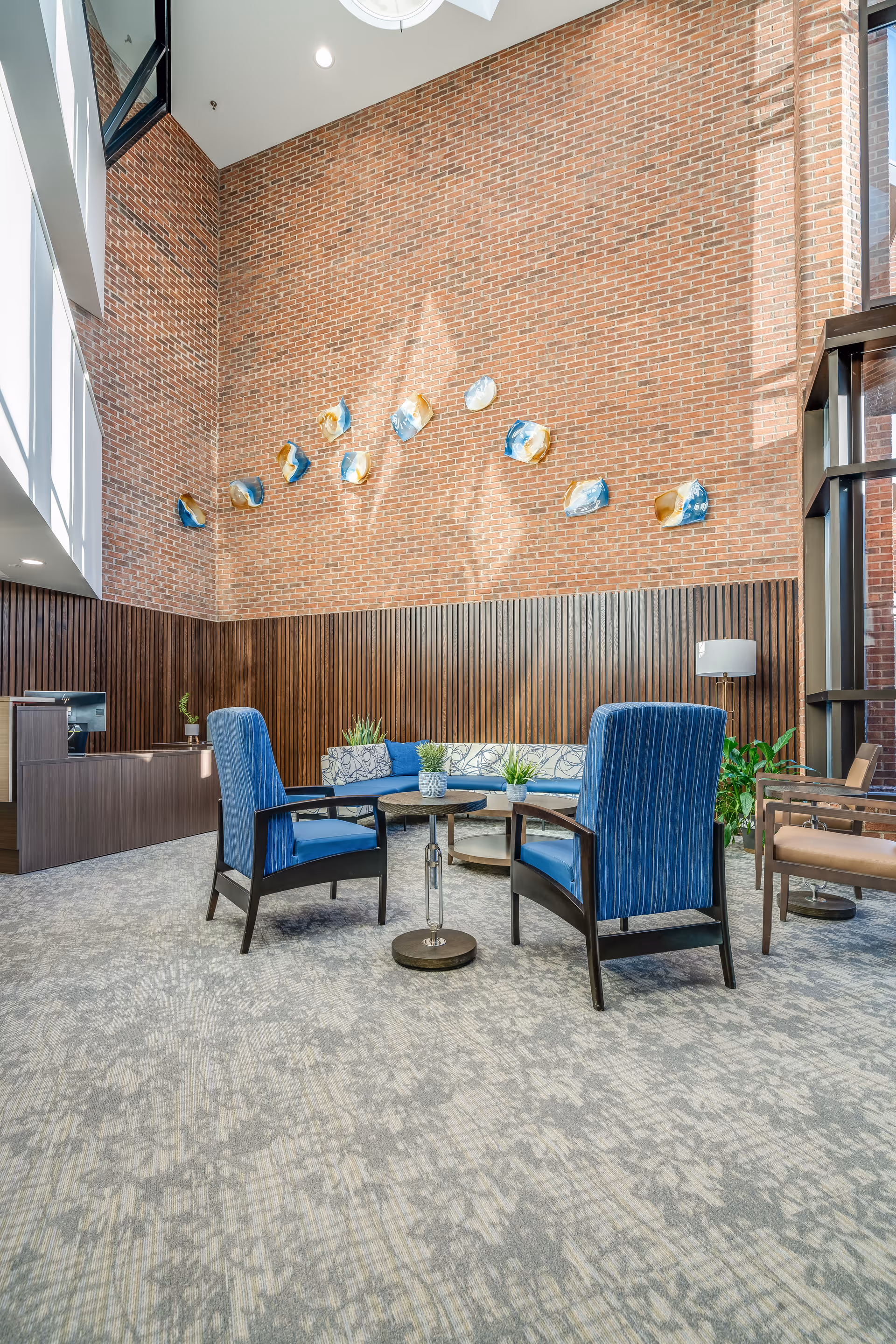A bright and spacious seating area with high ceilings and a large brick wall adorned with decorative art pieces. The room features two blue upholstered armchairs, a round table with small potted plants, a blue cushioned bench, and additional seating. There is a wooden reception desk on the left and large windows on the right letting in natural light.