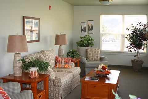 A cozy living room with patterned sofa and armchairs, wooden coffee and side tables, table lamps, and potted plants near a sunlit window.