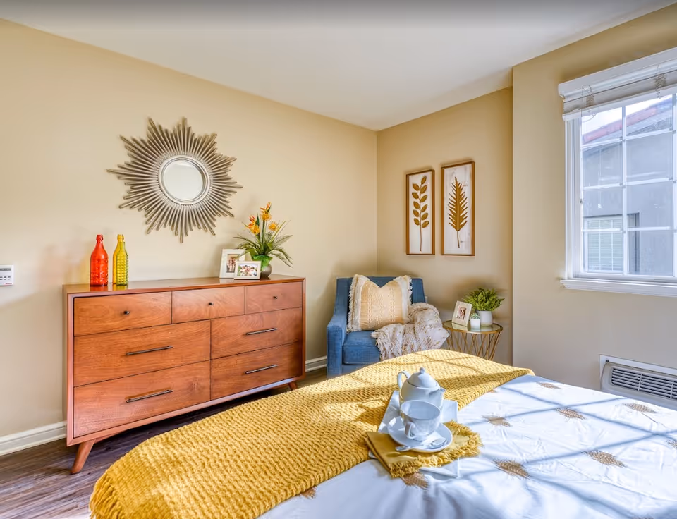 Sunlit bedroom with a wooden dresser, decorative sunburst mirror, blue armchair, and a bed with a yellow throw.
