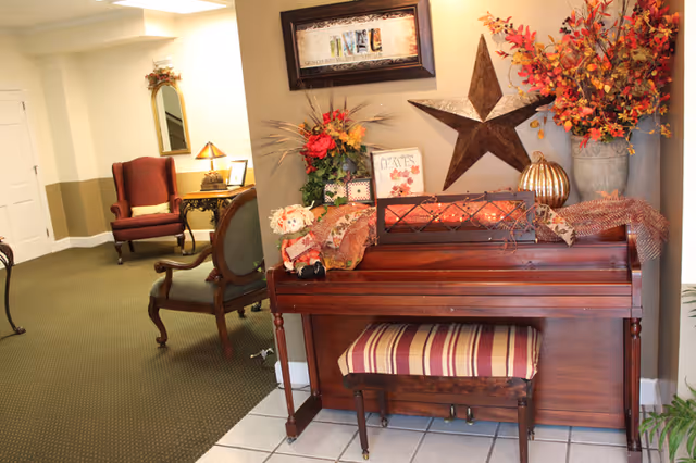 An interior corner of a senior living facility featuring a wooden piano with a striped cushioned bench. The piano is decorated with autumn-themed items including a metal star, a pumpkin, a floral arrangement, and a small scarecrow doll. In the background, there are two upholstered chairs, a small table with a lamp, and a mirror on the wall.