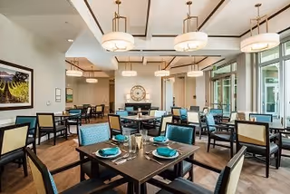 A spacious and well-lit dining room in a senior living facility with multiple tables set with teal napkins, plates, and glassware. The room features modern light fixtures hanging from the ceiling, large windows allowing natural light, and a decorative clock on the far wall.