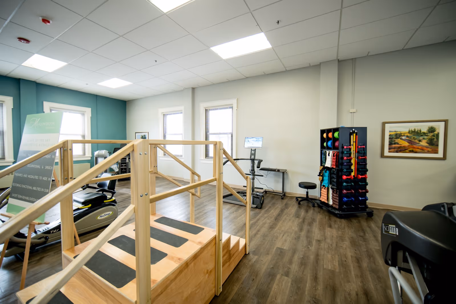 Well-lit therapy/fitness room with wooden exercise steps, cardio equipment, and a rack of colorful weights.