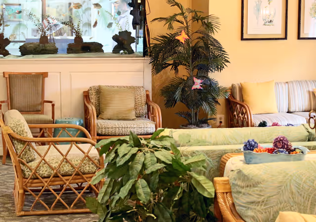 A cozy living room area in an assisted living facility featuring wicker and cushioned chairs, a striped sofa with yellow pillows, a large indoor plant decorated with butterfly ornaments, framed botanical artwork on the wall, and a large aquarium with fish in the background.