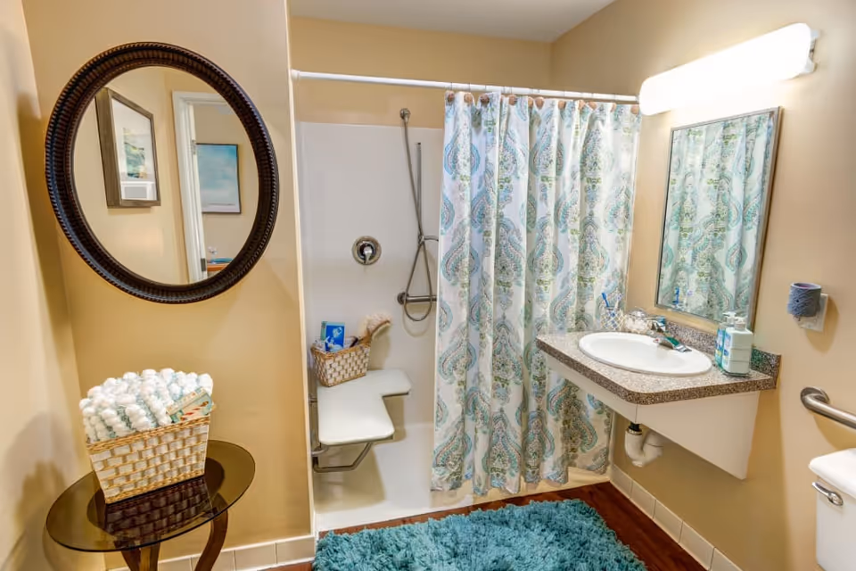 A clean and well-lit bathroom featuring a shower with a patterned curtain and a fold-down seat, a wall-mounted sink with a mirror above it, a toilet partially visible on the right, a round decorative mirror on the left wall, a small table holding a basket of rolled towels, and a blue textured bath mat on the floor.