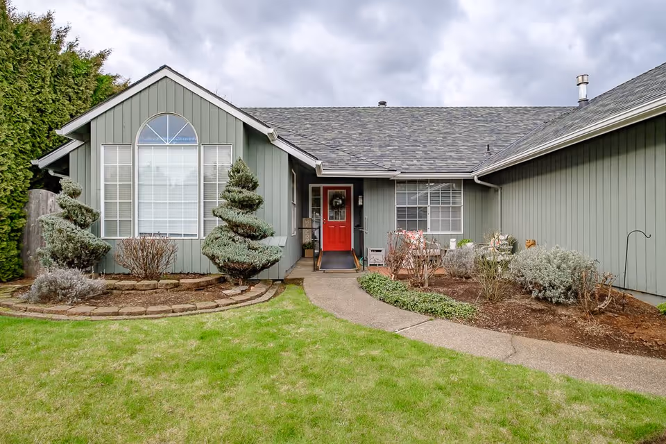 Front exterior view of a single-story building with gray siding and a red front door. The entrance is flanked by two spiral-shaped evergreen shrubs and other landscaping with a green lawn and a concrete pathway leading to the door. The sky is cloudy.