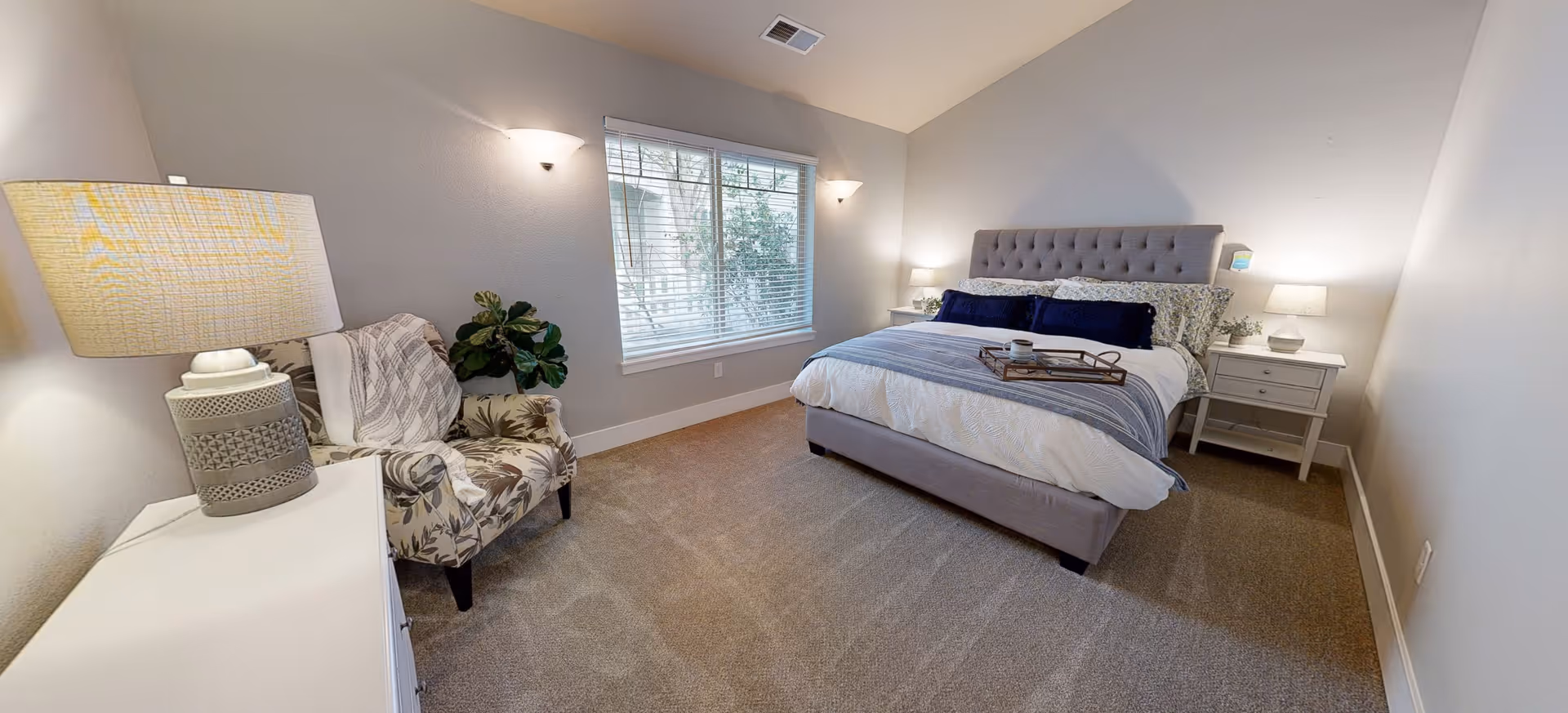 A cozy bedroom with a large bed featuring a tufted headboard, white and blue bedding, and a tray on top. There are two white nightstands with lamps on either side of the bed. A window with blinds lets in natural light. A patterned armchair with a throw blanket is placed near a white dresser with a decorative lamp. The walls are painted light gray and the floor is carpeted.