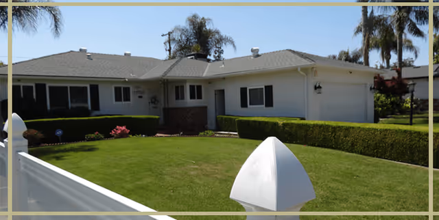 Single-story residential building with a well-maintained green lawn, trimmed hedges, and palm trees in the background under a clear blue sky.