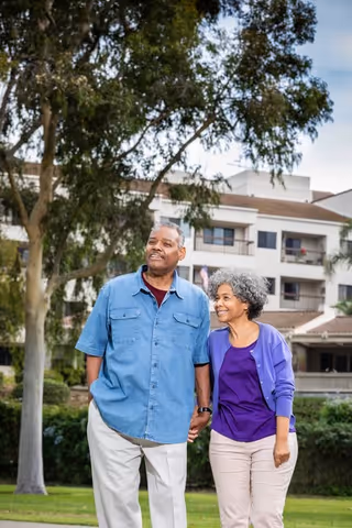 An elderly couple holding hands and walking outside in a garden area with trees and a multi-story building in the background.