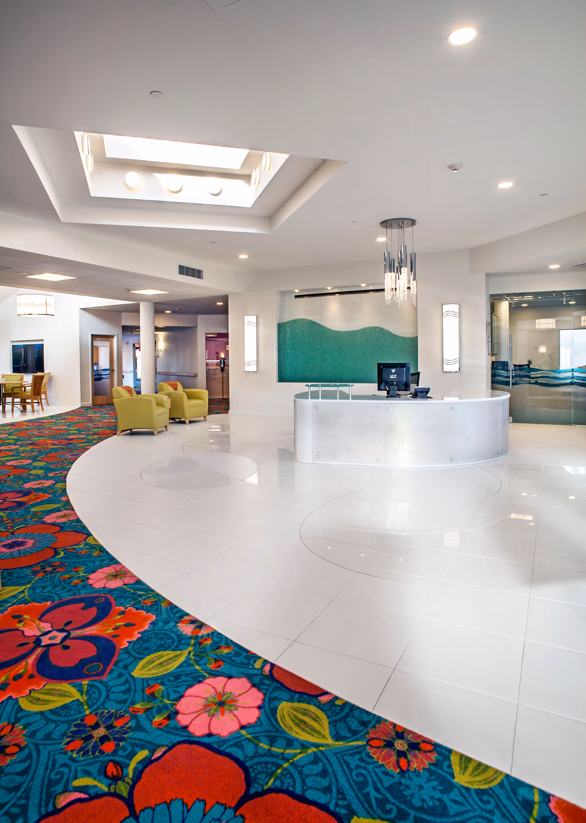 Bright and spacious reception area with a curved white desk, a computer on the desk, modern hanging light fixtures, and a large skylight above. The floor features a combination of white tiles and a colorful floral carpet. There are green armchairs and tables in the background near the entrance to other rooms.