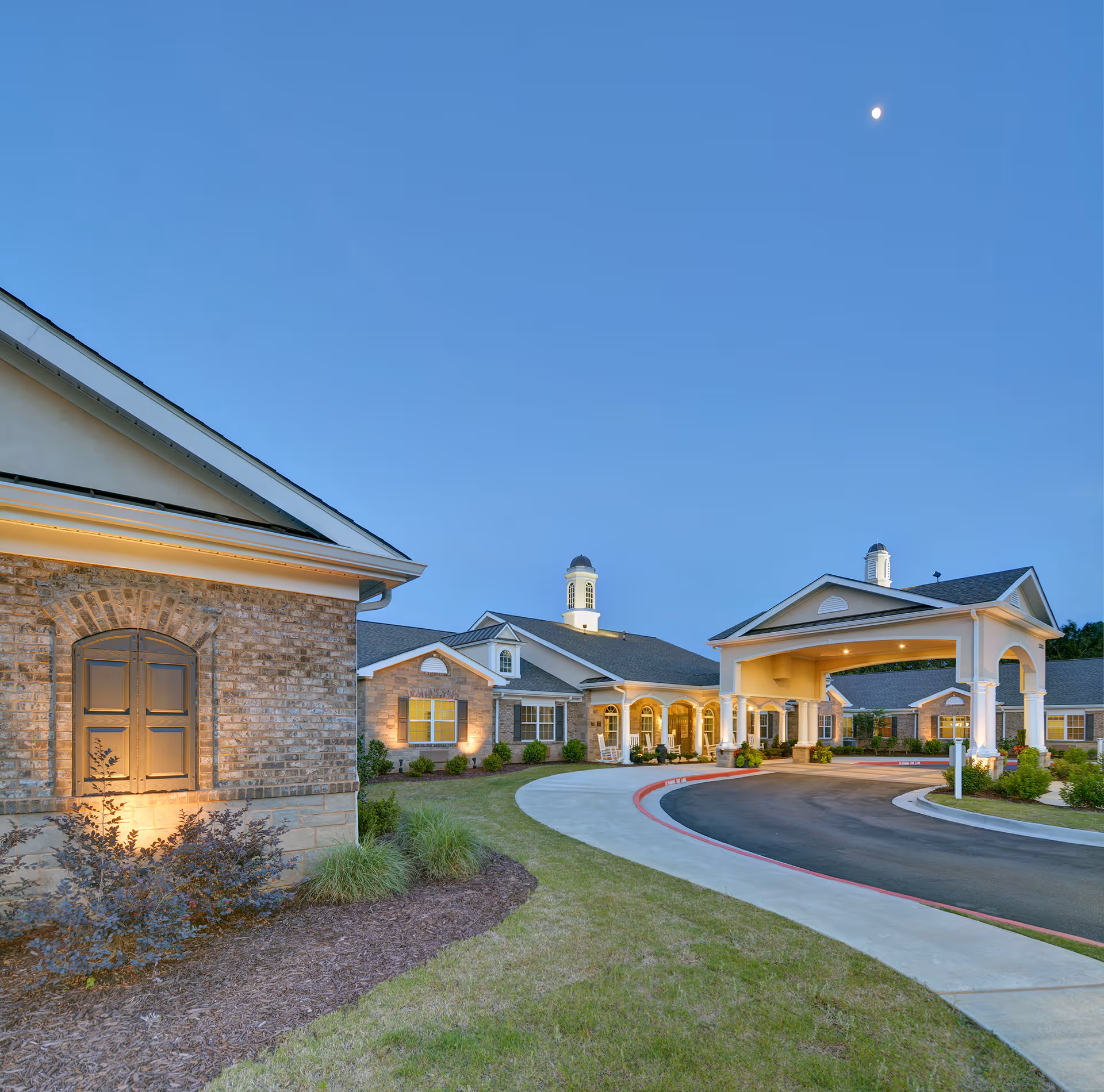 Exterior view of Benton Manor Of Stockbridge senior living facility at dusk, showing a curved driveway leading to a covered entrance with columns, well-maintained landscaping, and a clear sky with the moon visible.